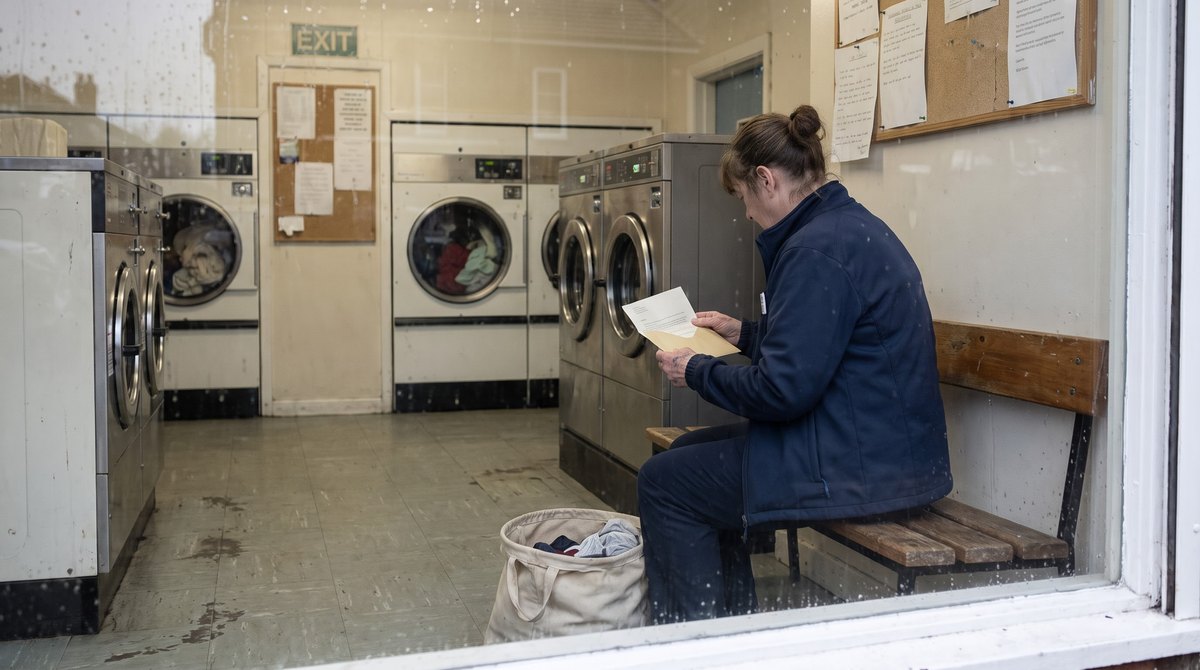 A launderette on a grey weekday afternoon, a woman in a supermarket uniform sitting on the bench with an opened letter held in both hands and a half-filled…