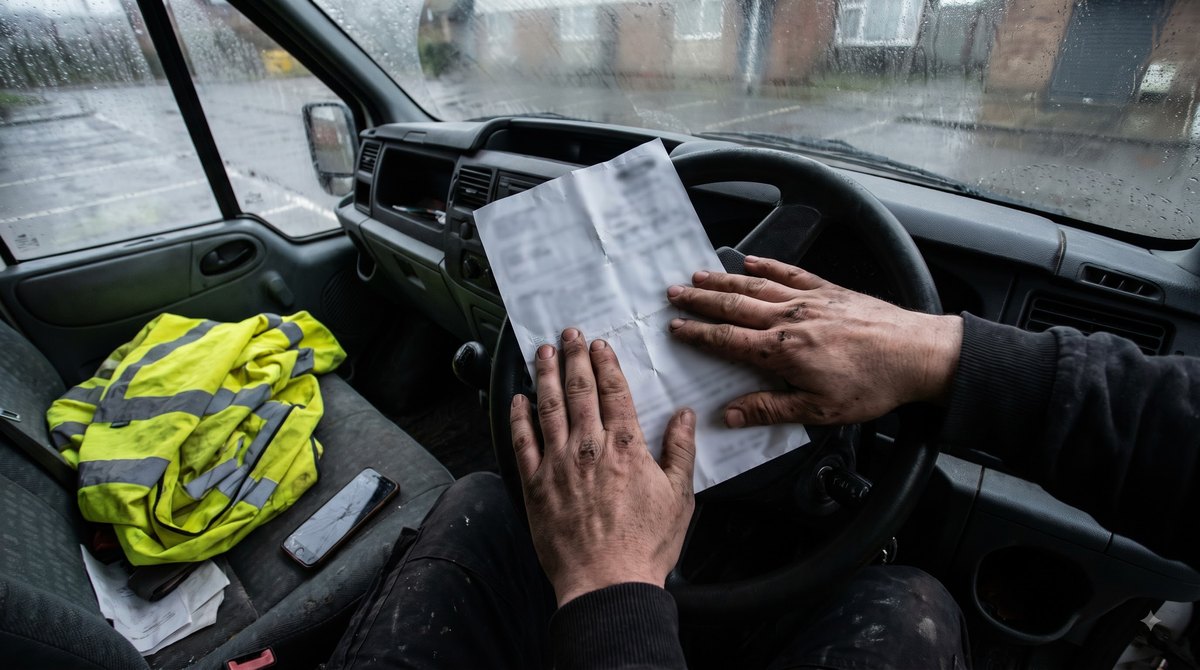 The cab of a builder's van at the end of a wet afternoon, two work-grubby hands smoothing a crumpled payslip flat against the steering wheel, a folded…